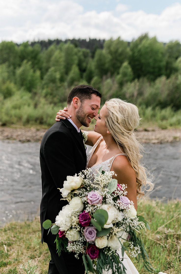 A bride in a lace wedding dress holding a bouquet, and a groom in a suit, smile at each other and lean in for a kiss, while standing outside, standing in front of a river and forest.