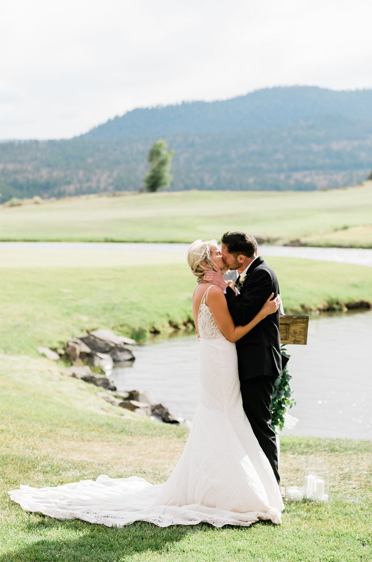 A bride in a lace wedding dress and a groom in a suit kiss outdoors by a pond, with hills in the background.
