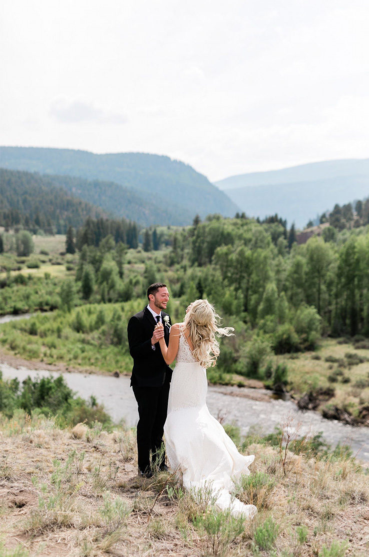 A bride in a lace wedding dress and a groom in a suit stand on a hill, facing each other, while holding hands, with a river, forest, and mountains in the background.