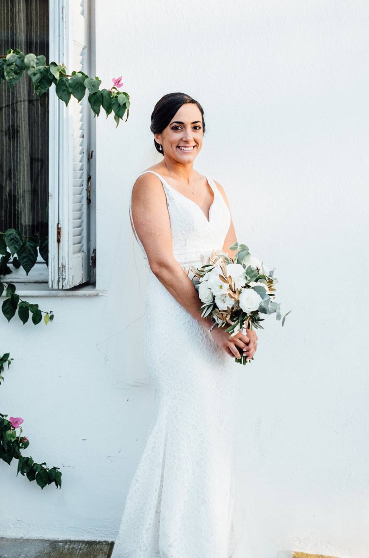 A bride in a sleeveless lace wedding dress holds a bouquet and smiles while standing outdoors in front of a wall and a window.