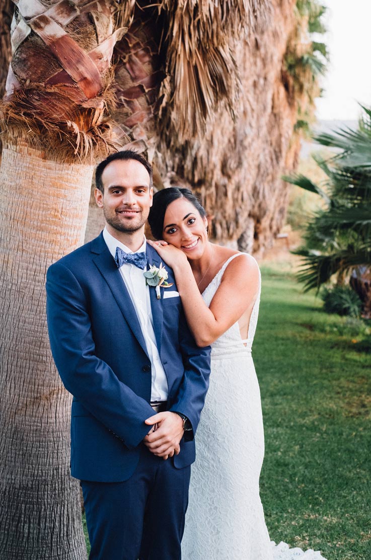 A bride in a sleeveless lace wedding dress rests her hands and head on the shoulder of a groom in a suit, both of them smiling, standing outside, in front of tall tree trunks.