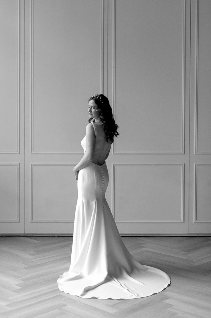 Back of bride in a fit and flare crepe gown with a buttoned low back, posing by a white wall in black and white.