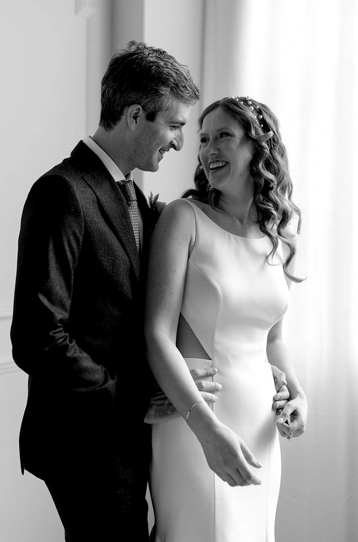 Groom embraces bride in a jewel neck crepe gown with side cutouts standing by a bright window, in black and white.
