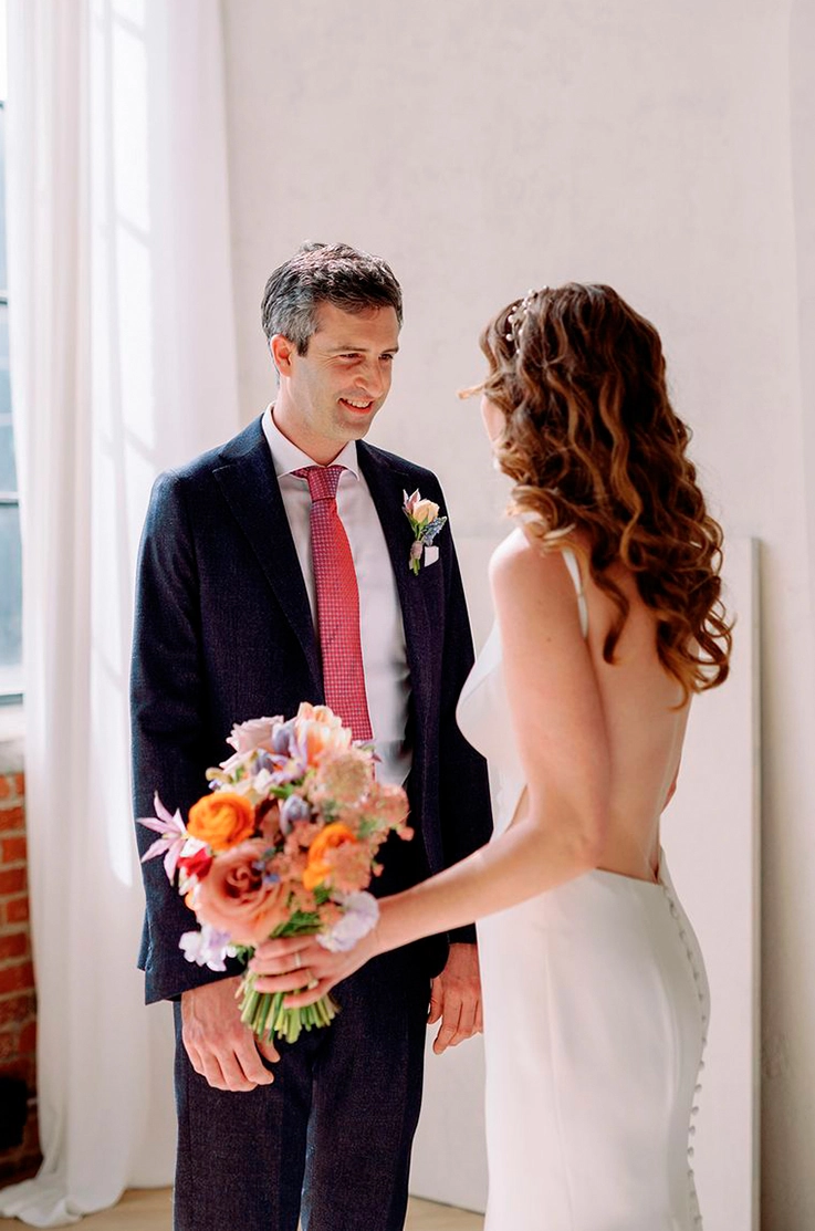 Bride in a sleek open back crepe gown with buttons, holds a bright colored bouquet and faces groom by a brick wall and window.