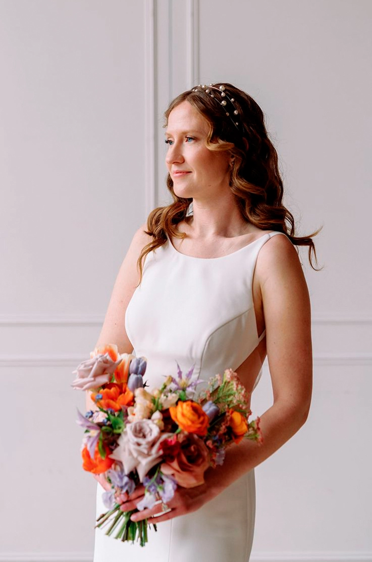 Close-up of bride smiling in a sleeveless crepe wedding dress with tulle side inserts, holding a colorful bouquet. 