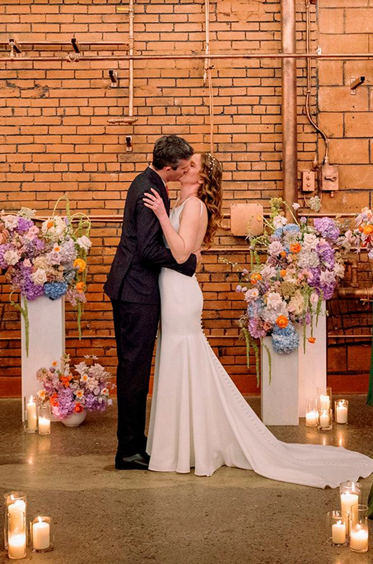 Bride in a sleek crepe gown with buttons and a low open back kisses groom at altar, decorated with flowers and candles.
