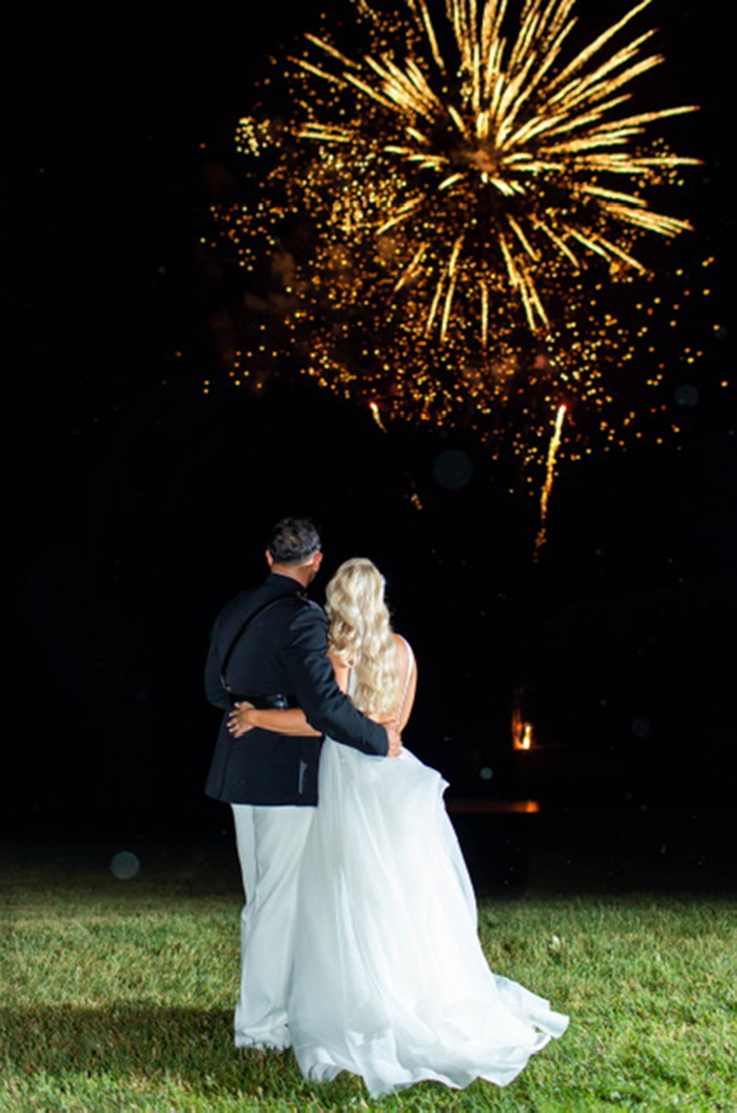 A bride in a sleeveless ball gown wedding dress and a groom in a marine dress uniform stand outside and watch fireworks burst against a dark sky.