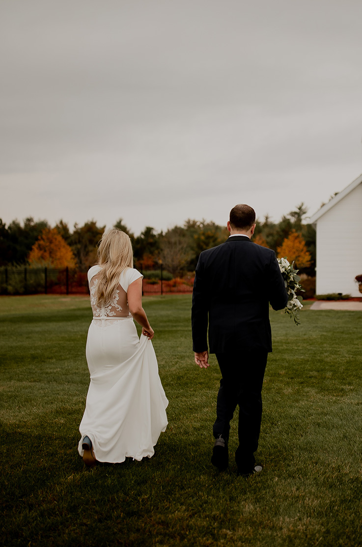 A bride in a statement back wedding dress and a groom in a suit holding a bouquet walk towards a building through a grass yard, with a forest in the background.