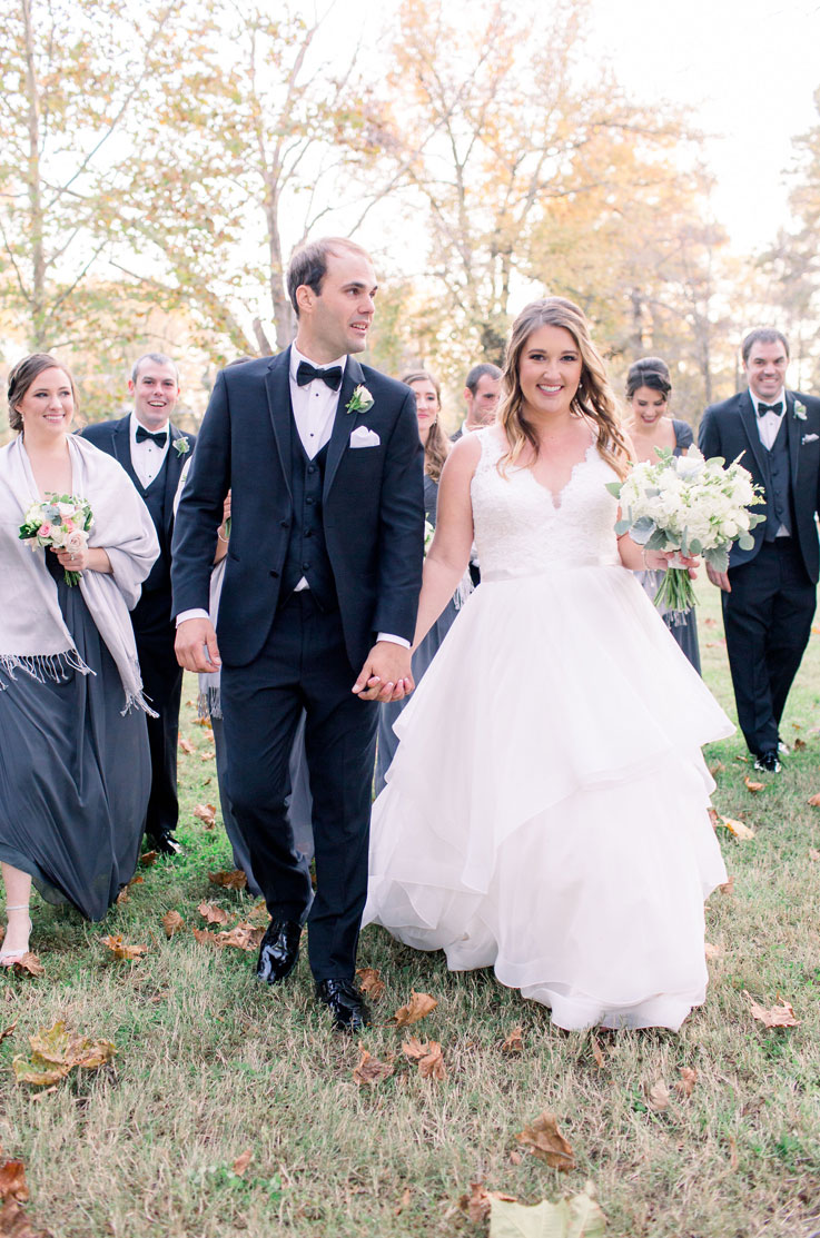 A bride in a lace and tulle wedding dress holds a bouquet and smiles while walking with a groom in a suit and their wedding party.