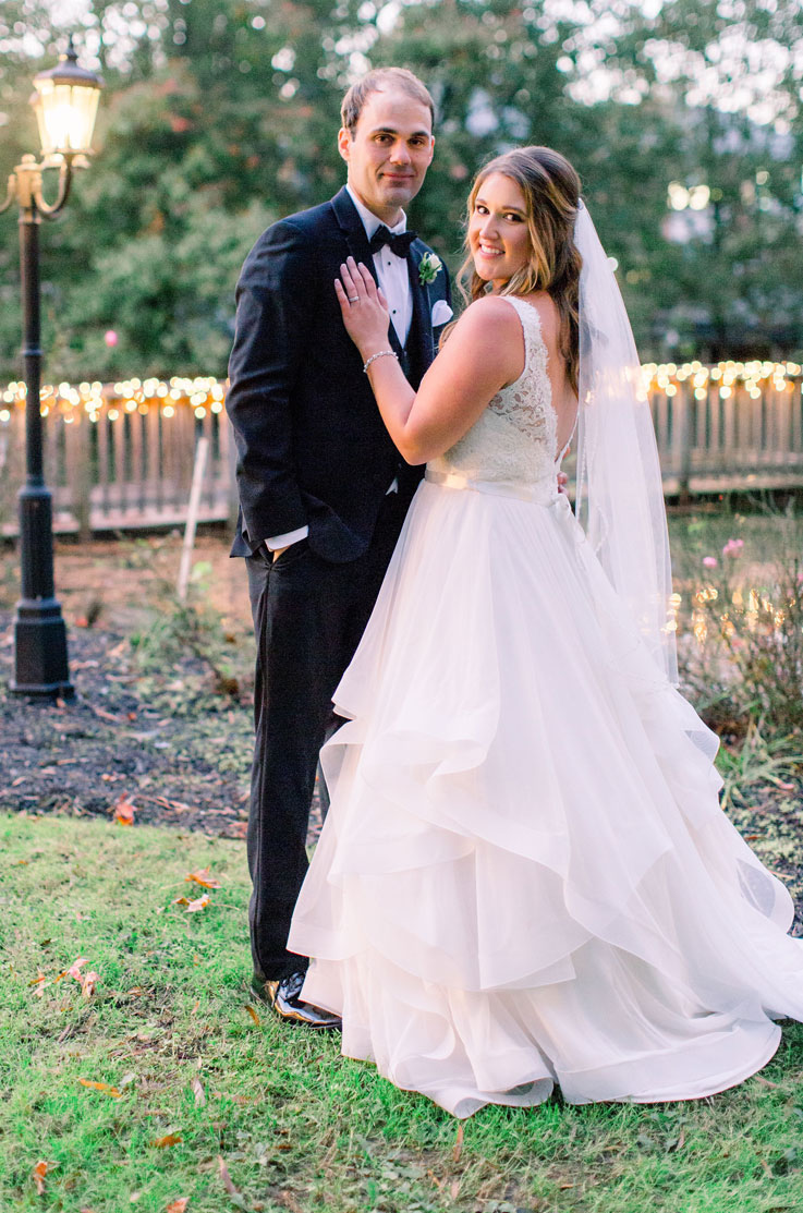 A bride in a lace and tulle wedding dress and a veil stands with a groom in a suit outside in a grass yard, in front of a fence lit with string lights.