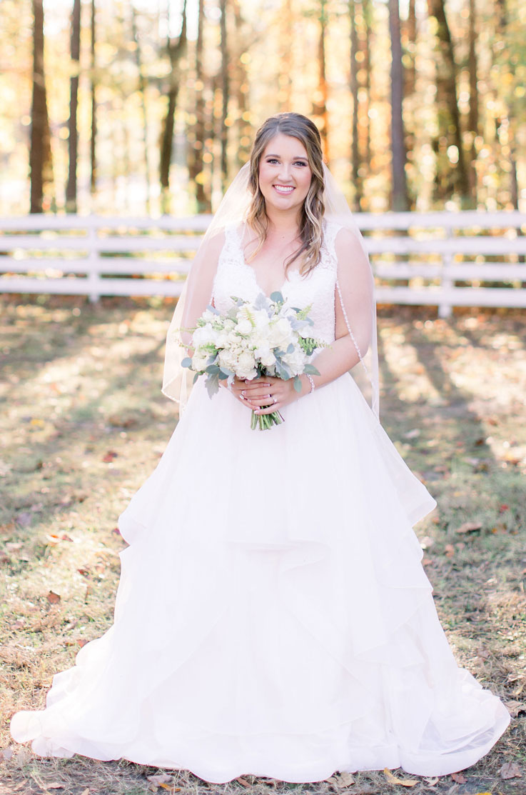 A bride in a lace and tulle wedding dress and a veil holds a bouquet while smiling and standing outside in front of trees and a fence.