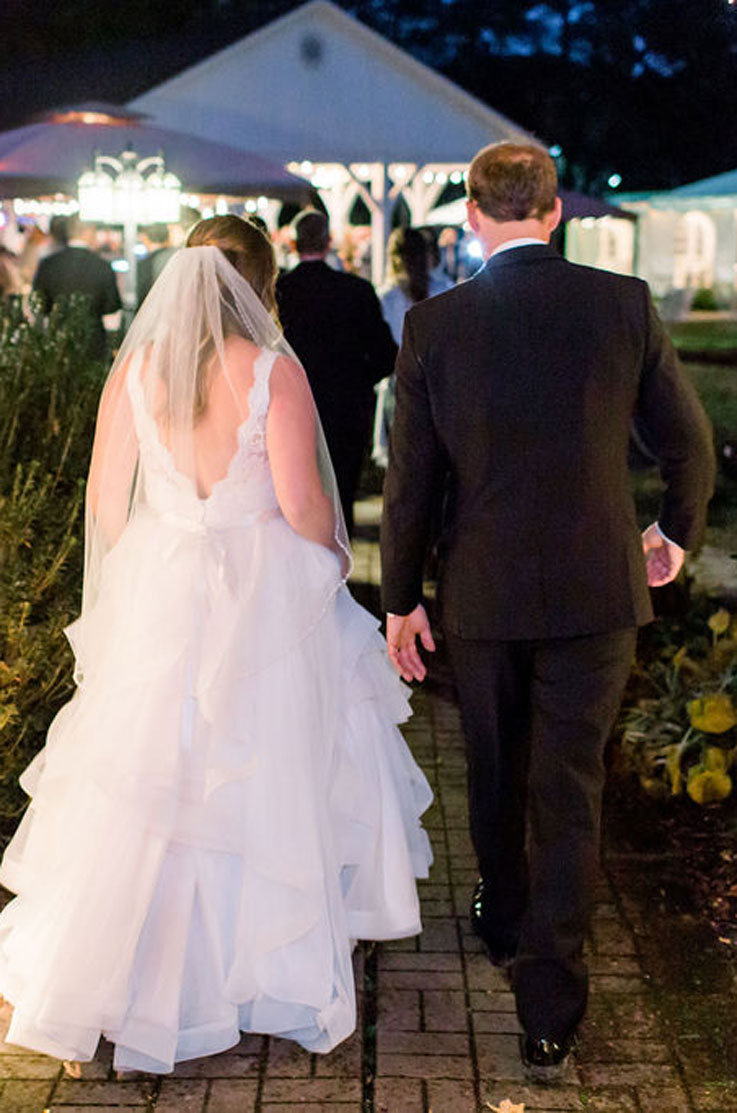 A bride in a lace and tulle wedding dress and veil walks down a path with a groom in a suit, towards an outdoor party.