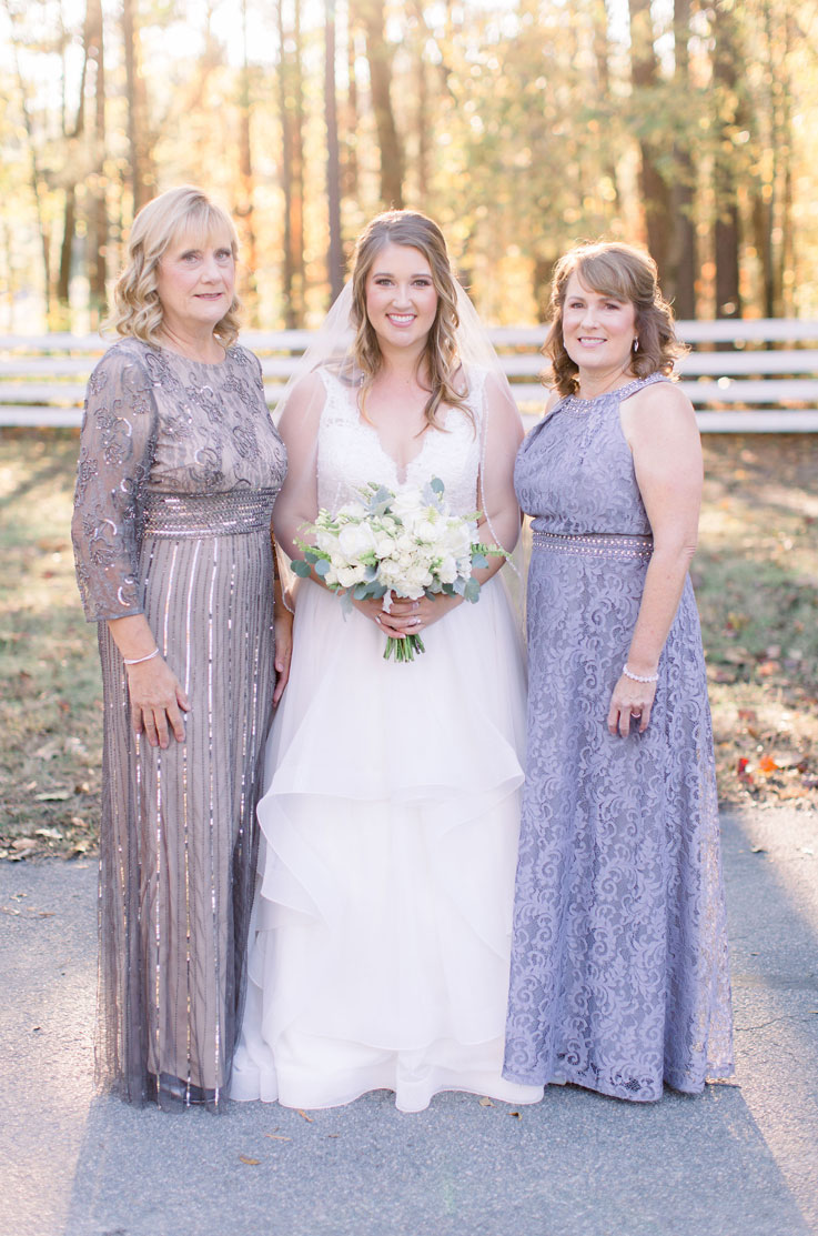 A bride in a lace and tulle wedding dress and a veil, holding a bouquet stands outside with two women in front of trees and a fence.