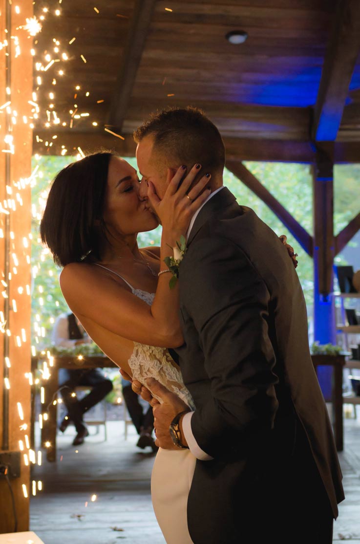 A bride in a fitted wedding dress and a groom in a suit kiss in a wooden pavilion with sparklers going off behind them.