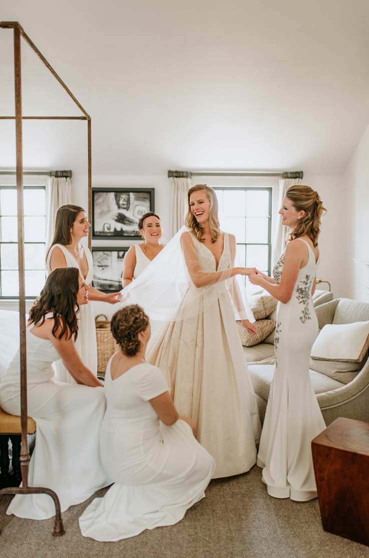 A bride in a V-neck ball gown wedding dress and veil is surrounded by her bridesmaids in a bedroom, while they all smile and look at the bride.