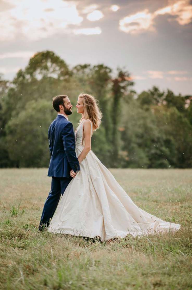 A bride in a ball gown wedding dress and a groom in a suit stand outside facing each other, in a field of grass with a forest behind them.