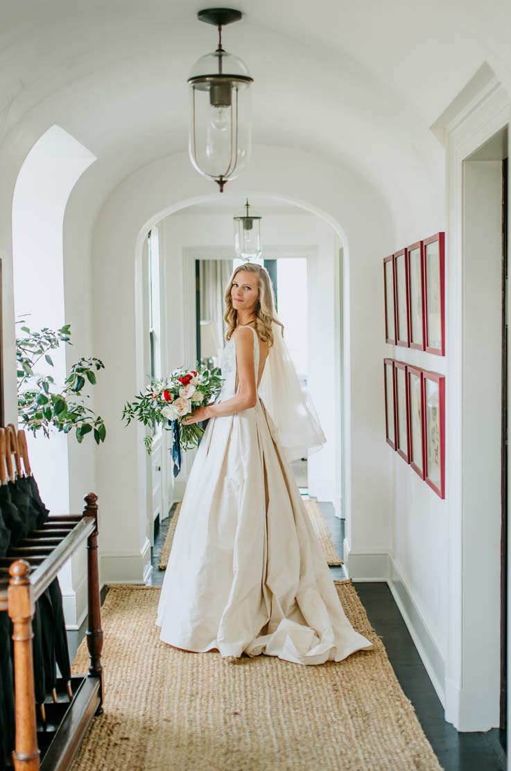A bride in a ball gown wedding dress holds a bouquet and stands in a hallway looking over her shoulder and smiling.