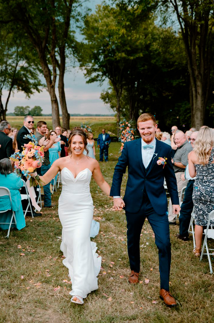 Bride in a strapless crepe fitted gown with a sweetheart neckline, walks down the aisle with groom and guests cheering. 