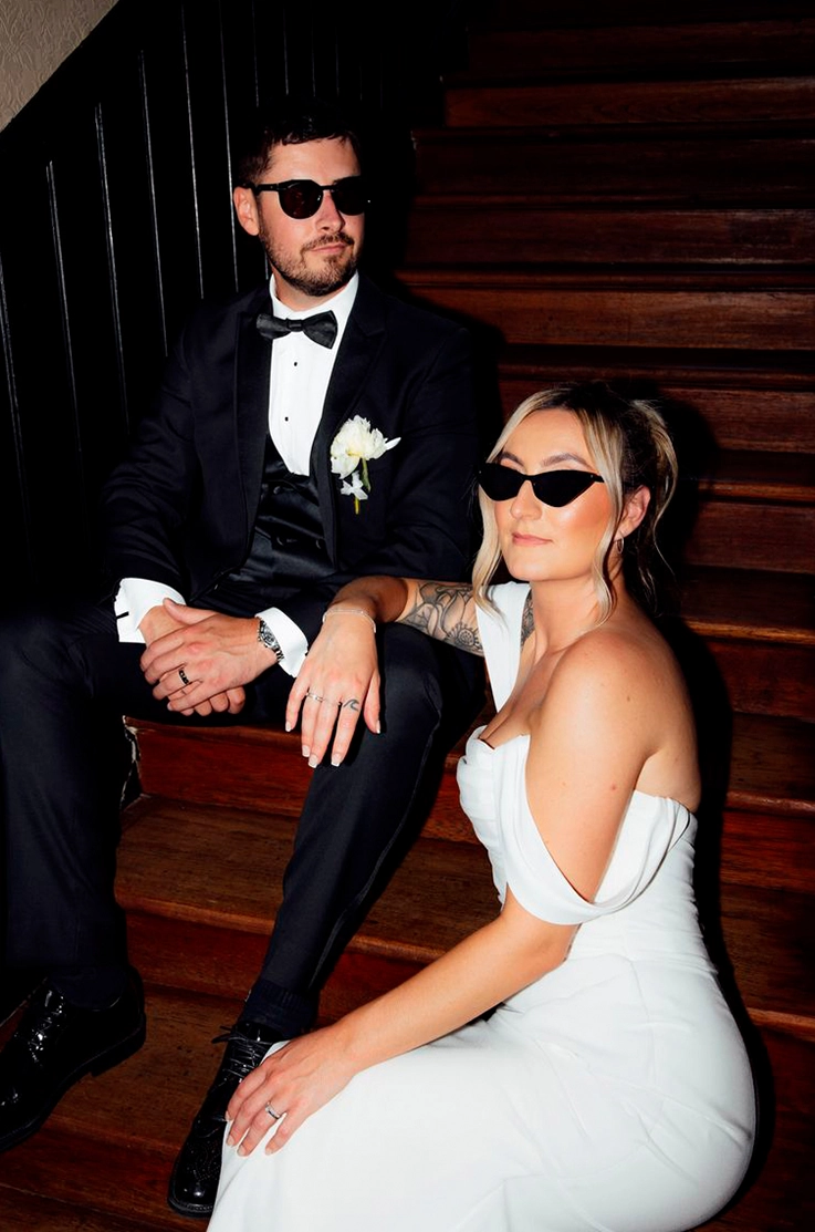 Bride in draped crepe bodice gown with sleeves, poses with groom in a tuxedo, sitting on steps and wearing sunglasses.