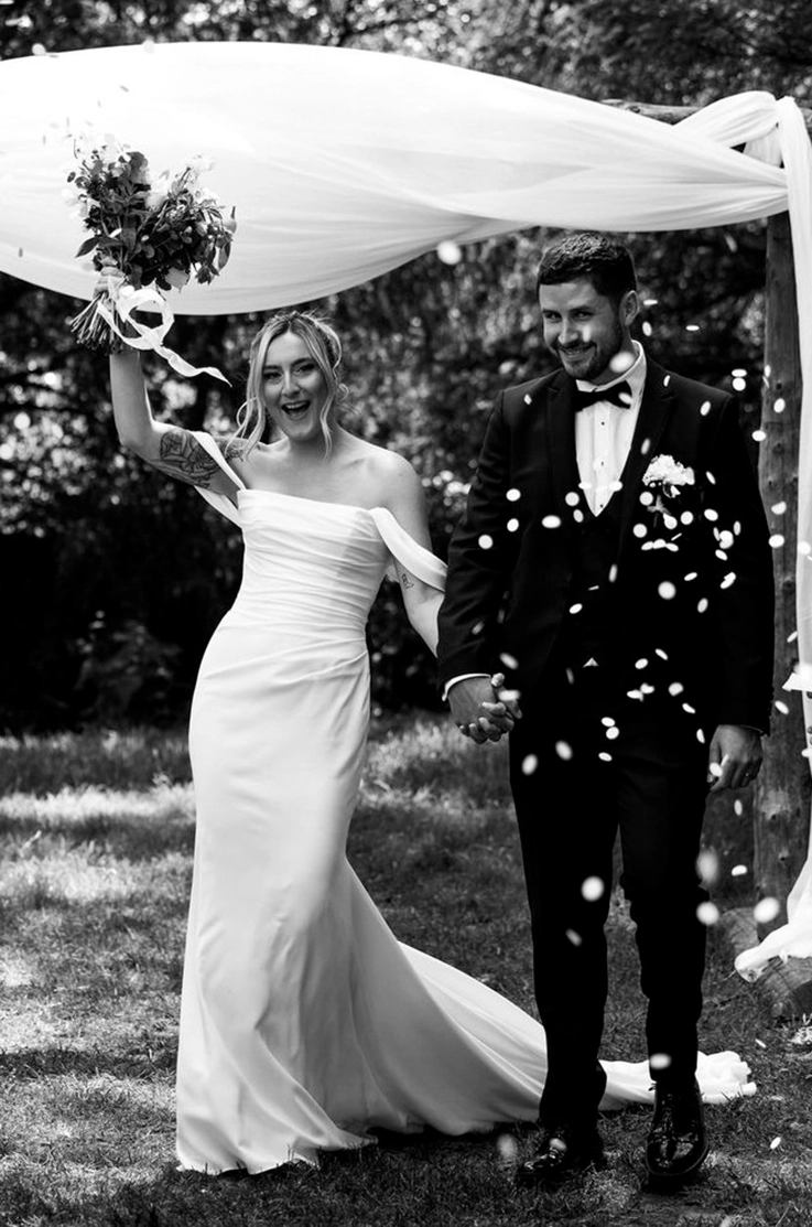 Bride in a fit and flare crepe gown with draped sleeves, holds up a bouquet celebrating with the groom at the altar. 