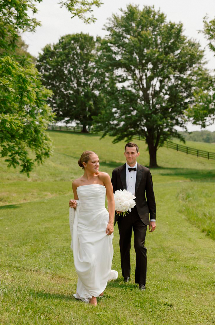 Bride in a strapless draped crepe gown walks in an open sunny meadow, alongside her groom in a tux holding a white bouquet.