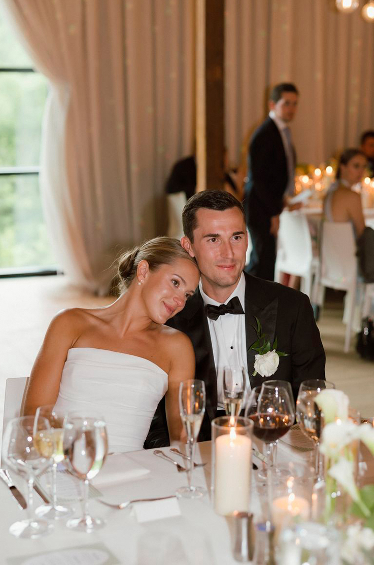 Bride in a strapless draped bodice gown, smiles next to groom in a tux, at a table by an open window and curtains.