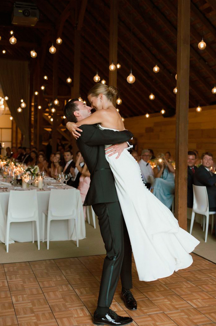 Groom picks up his bride in a strapless crepe wedding dress with a buttoned back, in front of guests under warm string lights.
