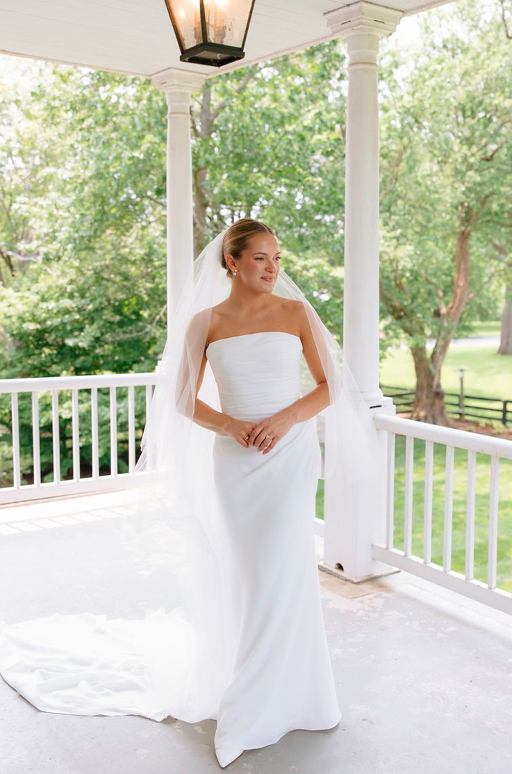 Bride in a strapless crepe fit and flare gown with a veil, poses on a wraparound white porch with two columns.
