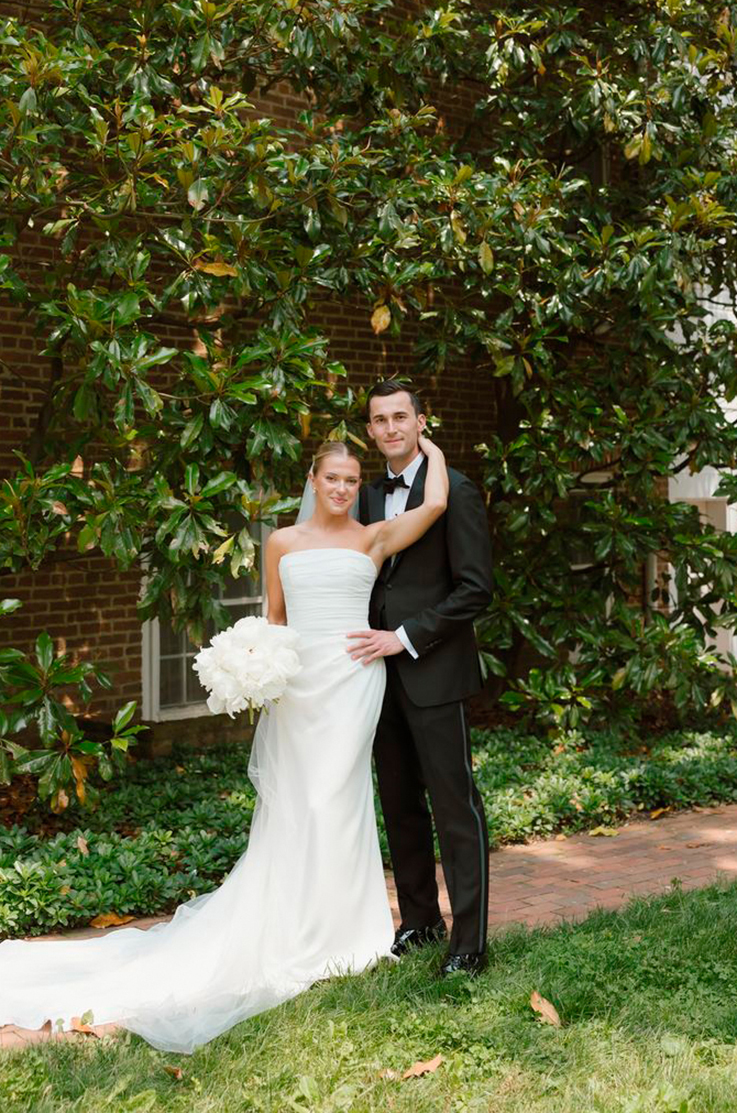 Bride in a draped crepe bodice gown with a veil, is holding a large white bouquet and poses with her groom by trees.