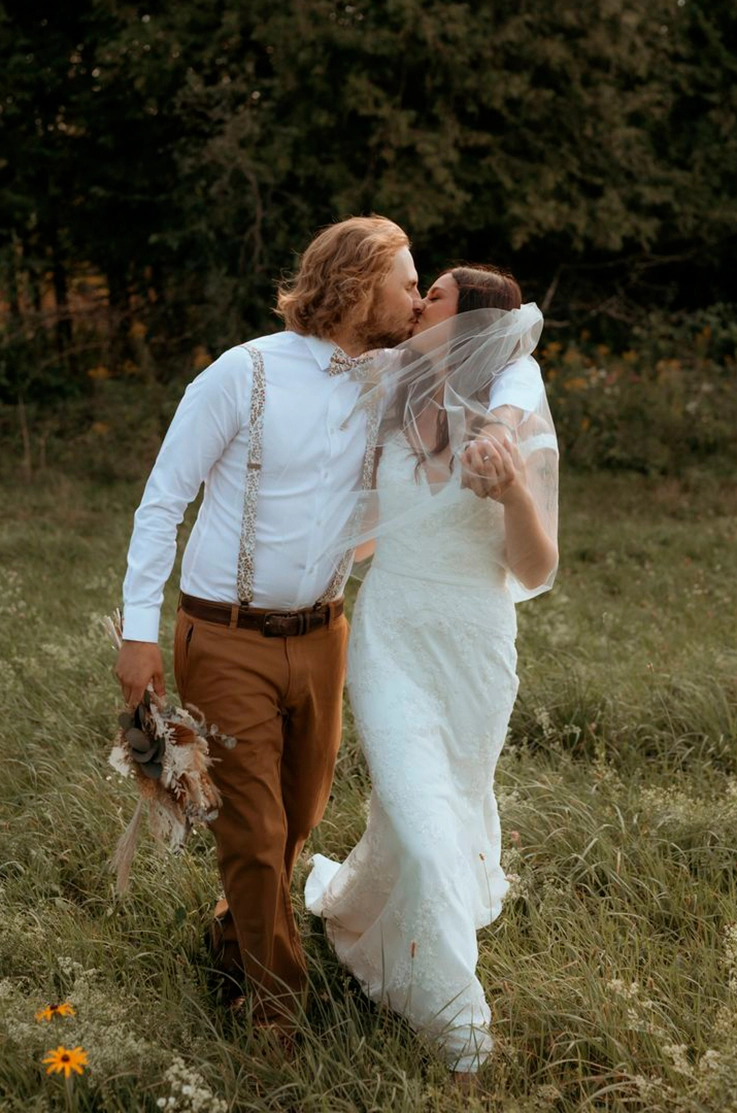 Bride in a lace fitted gown with a V-neckline and veil, walks beside her groom and they kiss in a wildflower meadow.