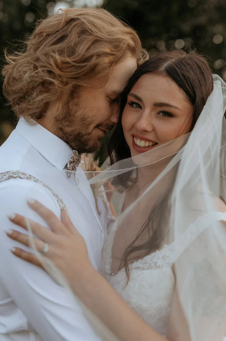 Close-up of groom embracing his bride, smiling in a lace V-neck wedding dress with a flowing veil outdoors.