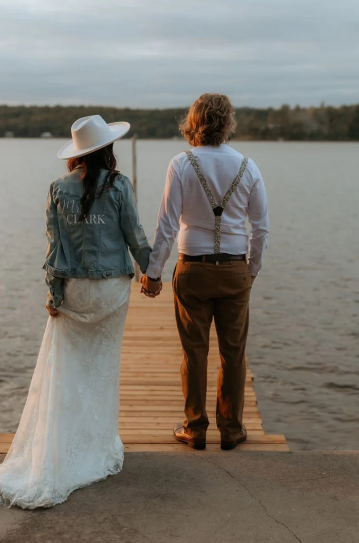 Back of bride in a fit and flare lace gown with a custom denim jacket overtop, holding hands with groom on a lakeside dock.