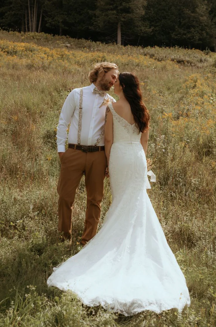 Bride in a V-shaped back lace gown poses alongside groom in an open meadow in the sunlight.