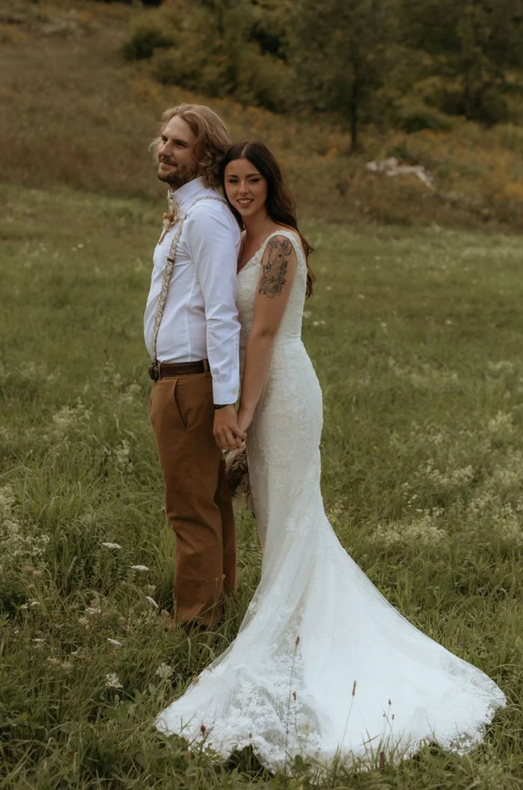 Bride in a V-neck off shoulder gown with a lace fit and flare skirt, poses beside the groom in an open meadow.