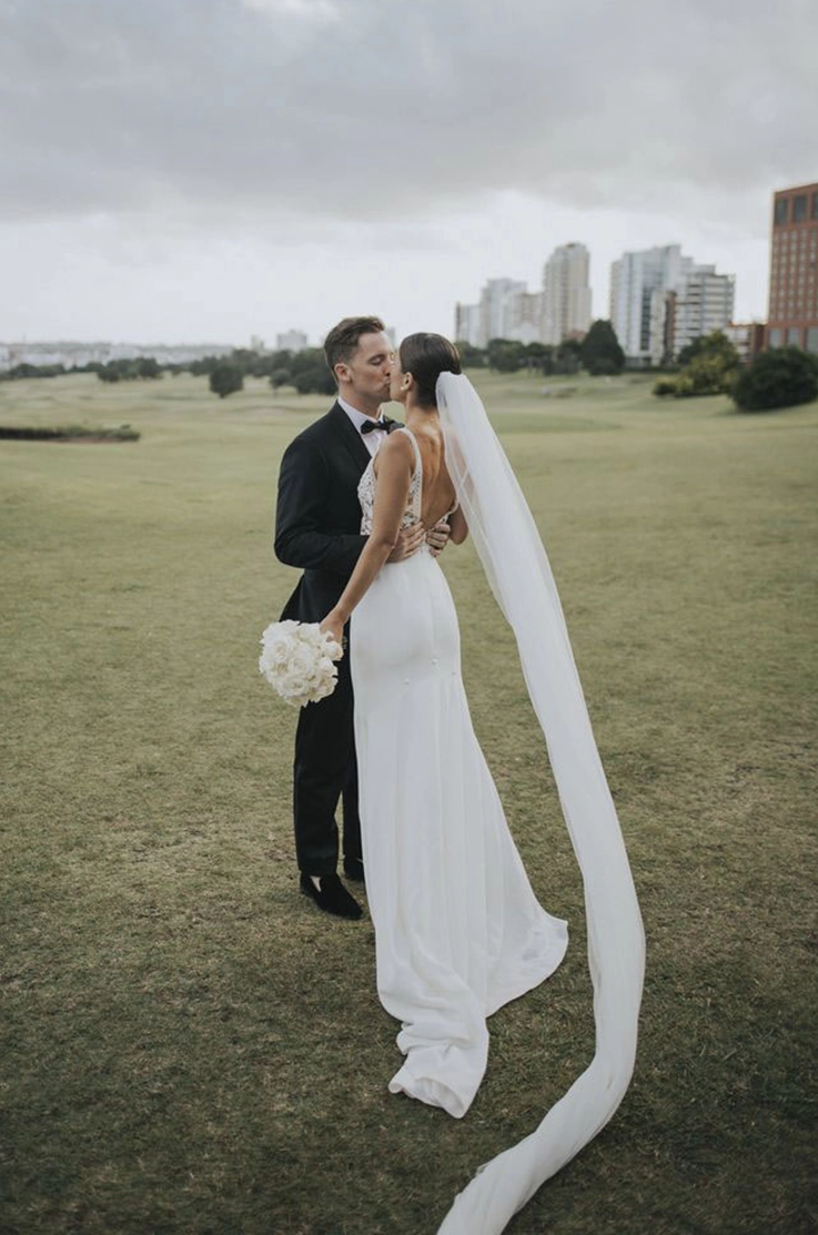 Groom kisses his bride in a lace bodice gown with a deep-V back, crepe skirt and a veil, standing in an open field.
