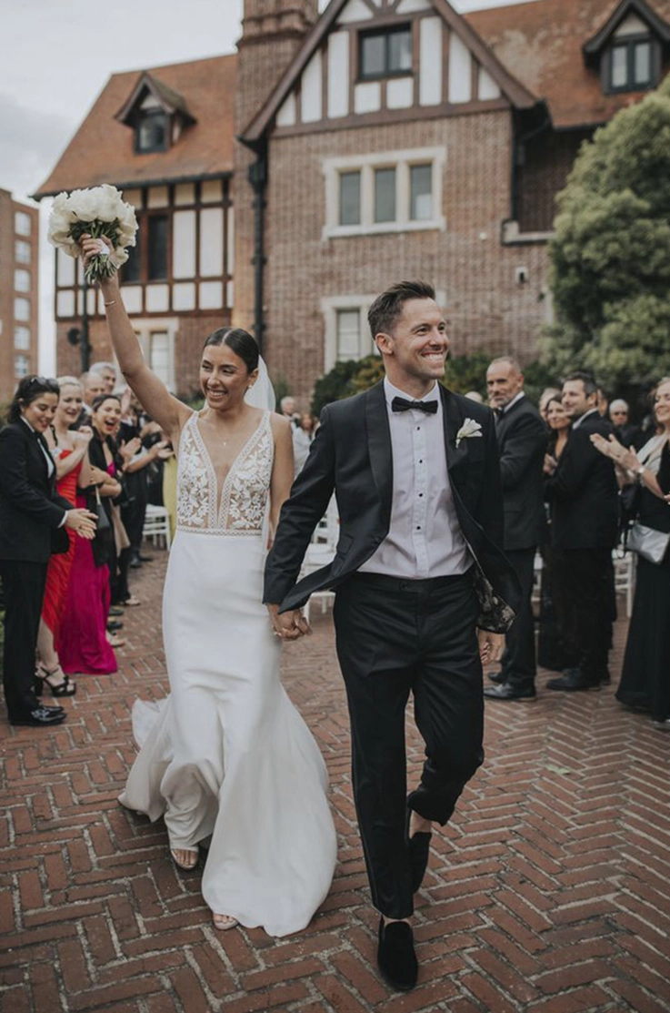 Bride in a V-neck lace and crepe wedding dress walks hand in hand with groom pasts guests outside the venue.