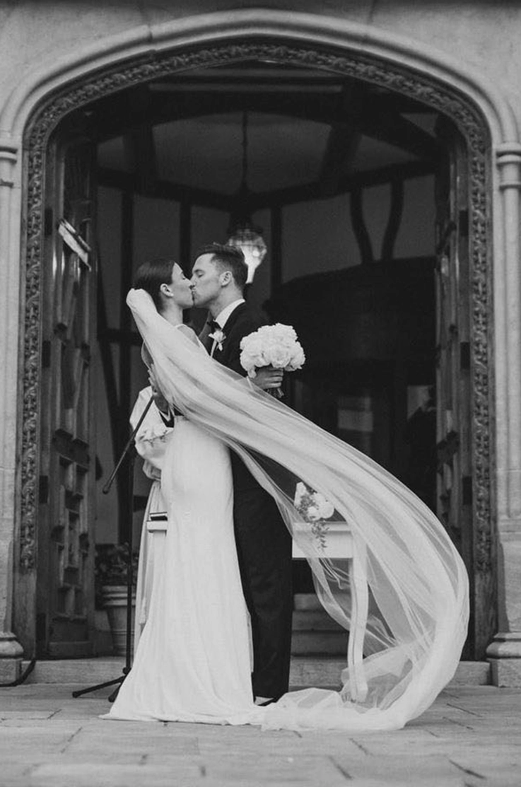 Bride in a sleeveless fit and flare gown kisses her groom, holding a bouquet in the doorway of the venue.