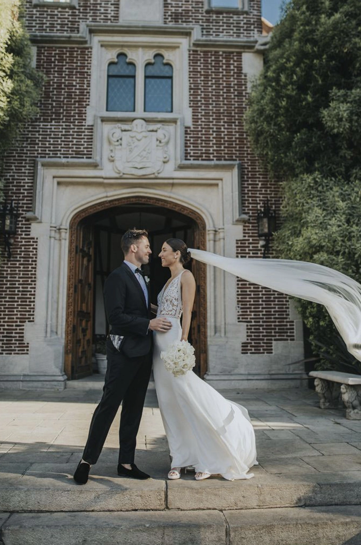 Bride in a V-neck lace gown with a crepe fit and flare skirt and veil, poses with her groom outside the venue entrance. 