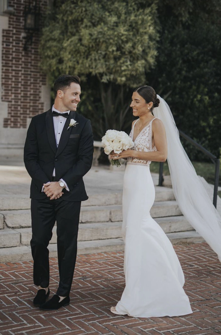 Bride in a lace bodice gown with a V-neck and crepe fit and flare skirt, smiles and is holding a bouquet beside the groom.