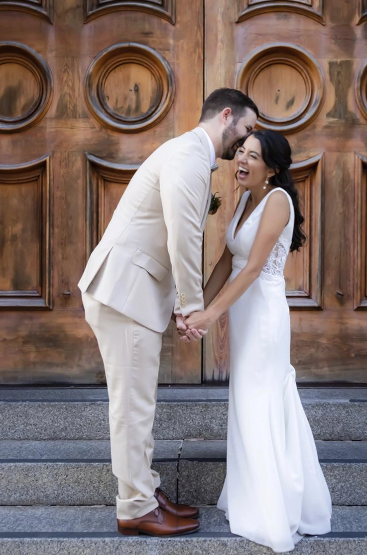Bride in a sleeveless crepe wedding dress with a V-neck and lace side details, laughs alongside groom at wooden double doors.