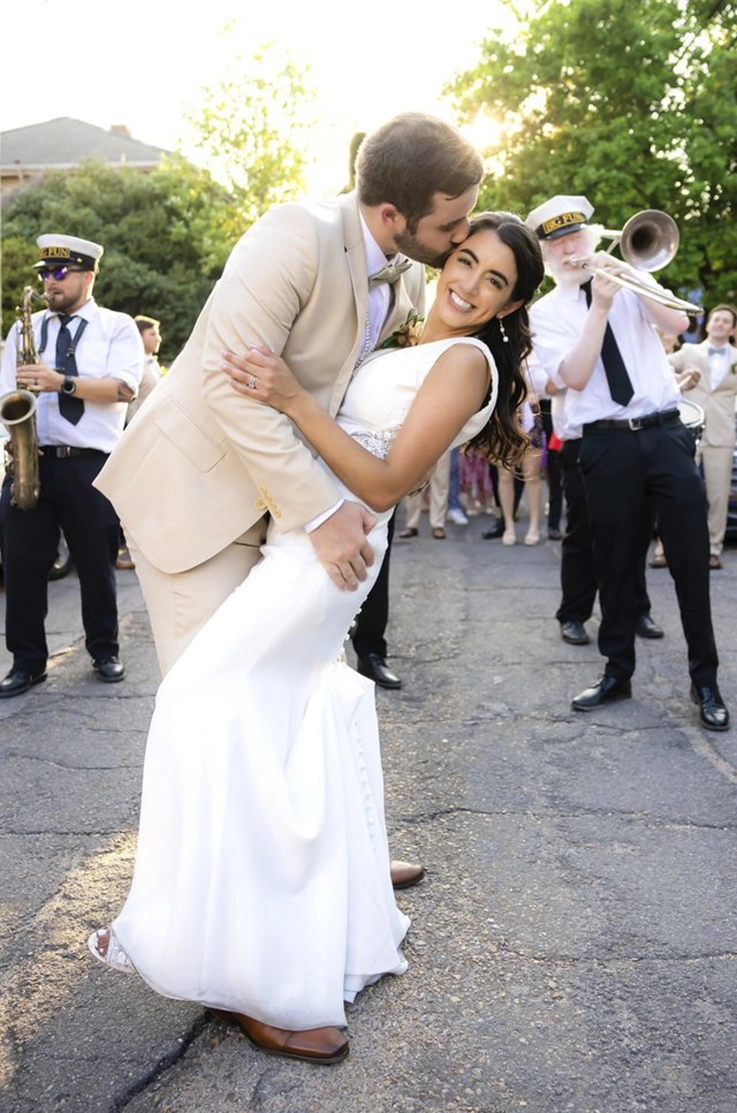 Groom embraces bride in a crepe bodice gown with buttons and a fit and flare skirt, in front of a brass band playing outdoors.