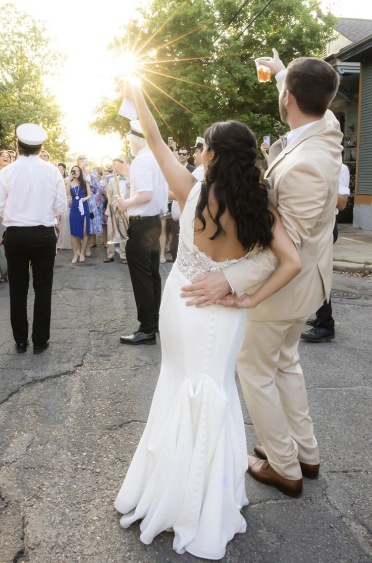 Back of bride in a crepe fit and flare gown with lace detail, stands alongside the groom as they raise drinks in their hands.