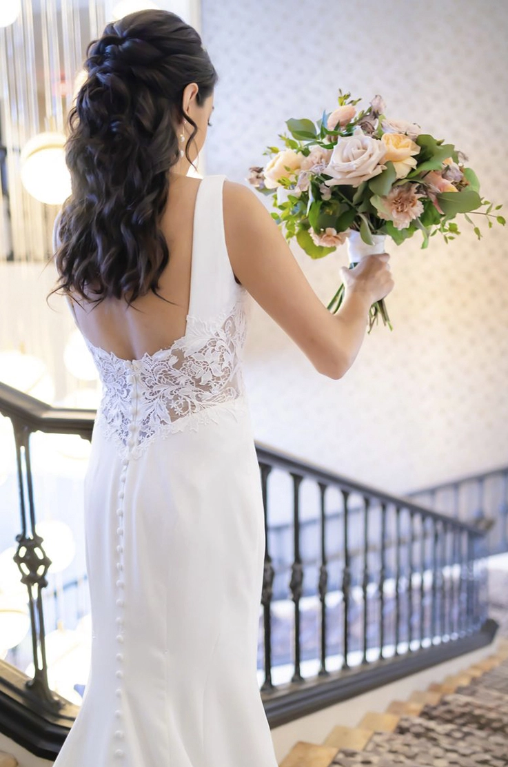 Close-up of bride in a lace and crepe gown with buttons down the back, is standing on a staircase and holding a bouquet.