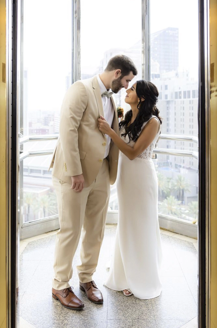 Bride in a lace and crepe wedding gown, poses and is facing her groom in a modern high-rise with tall windows.