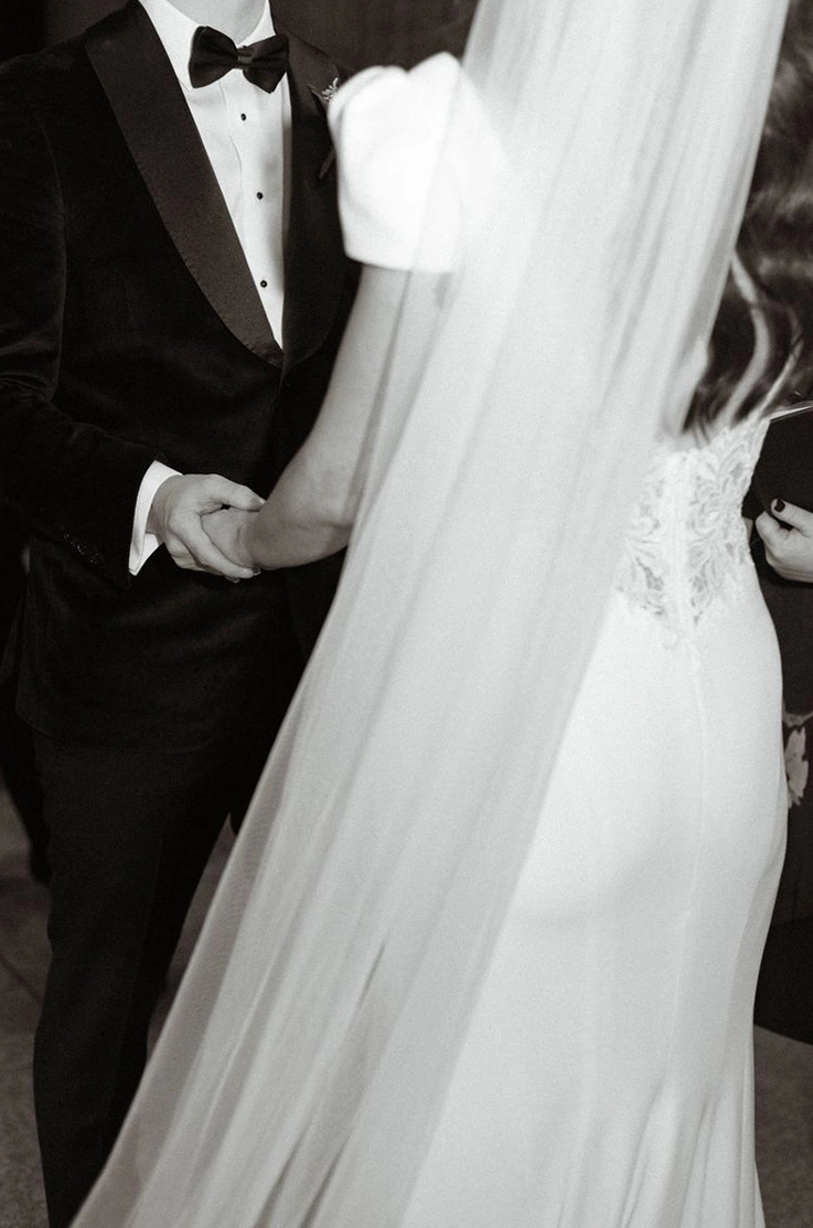 Close-up of bride in a crepe fit and flare wedding dress with lace back detail and a veil, faces groom in black and white.