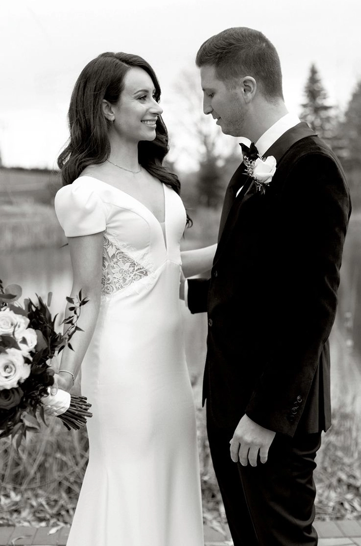 Bride in a lace and crepe gown with a V-neck and puffed cap sleeves, is holding a bouquet by a lake and poses next to her groom. 