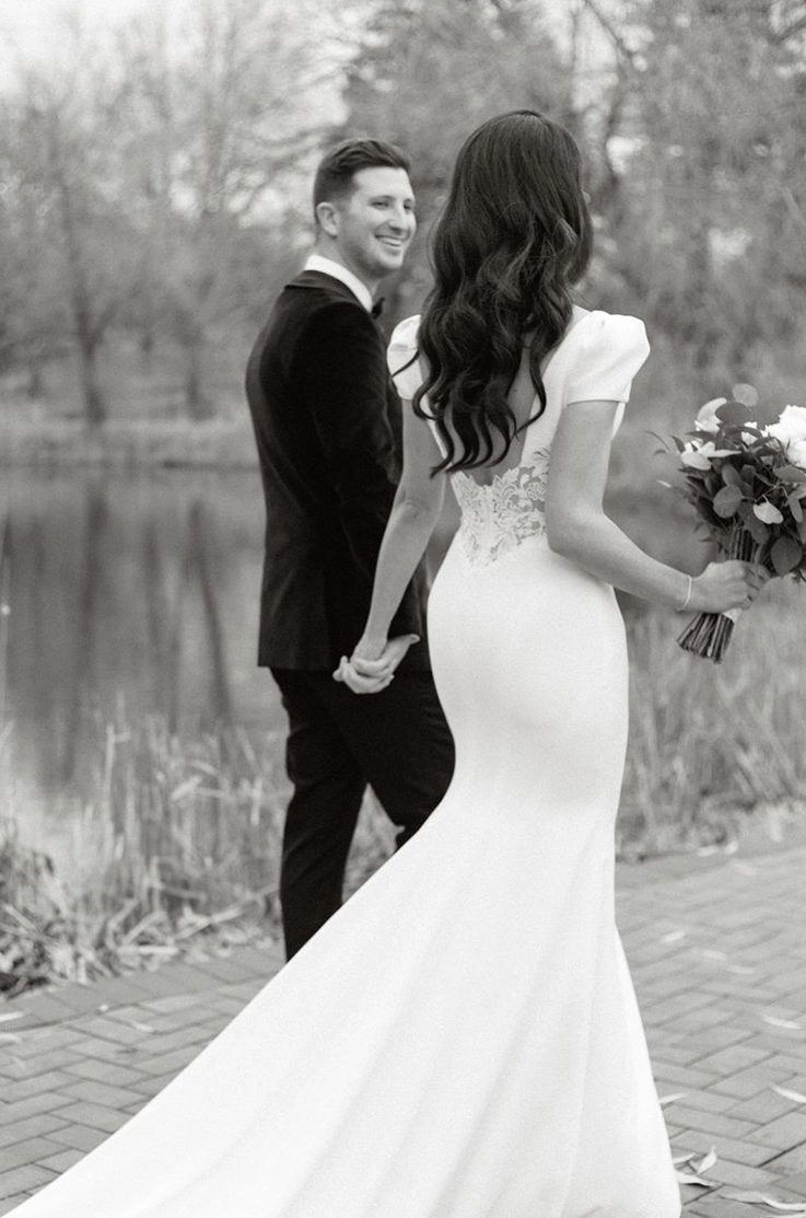 Bride in a crepe fit and flare gown with a lace low back, walks beside groom, along a lake in black and white. 