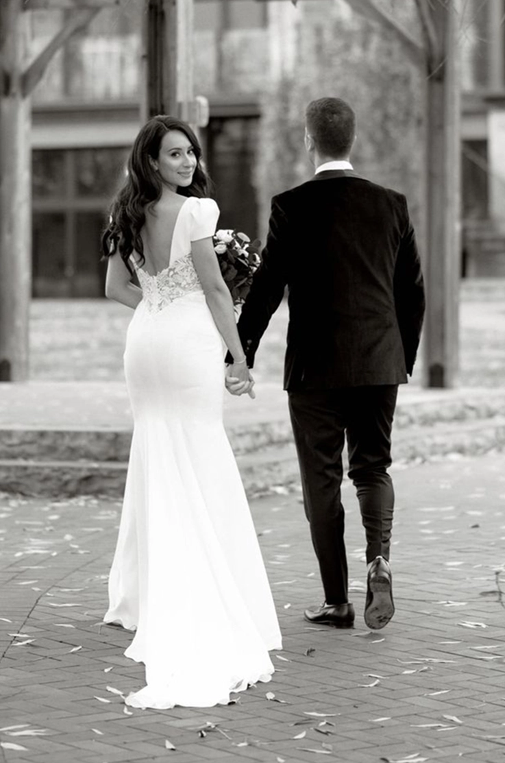 Bride in a cap sleeve crepe gown with lace detail and a low back, walks next to her groom outdoors looking over her shoulder.