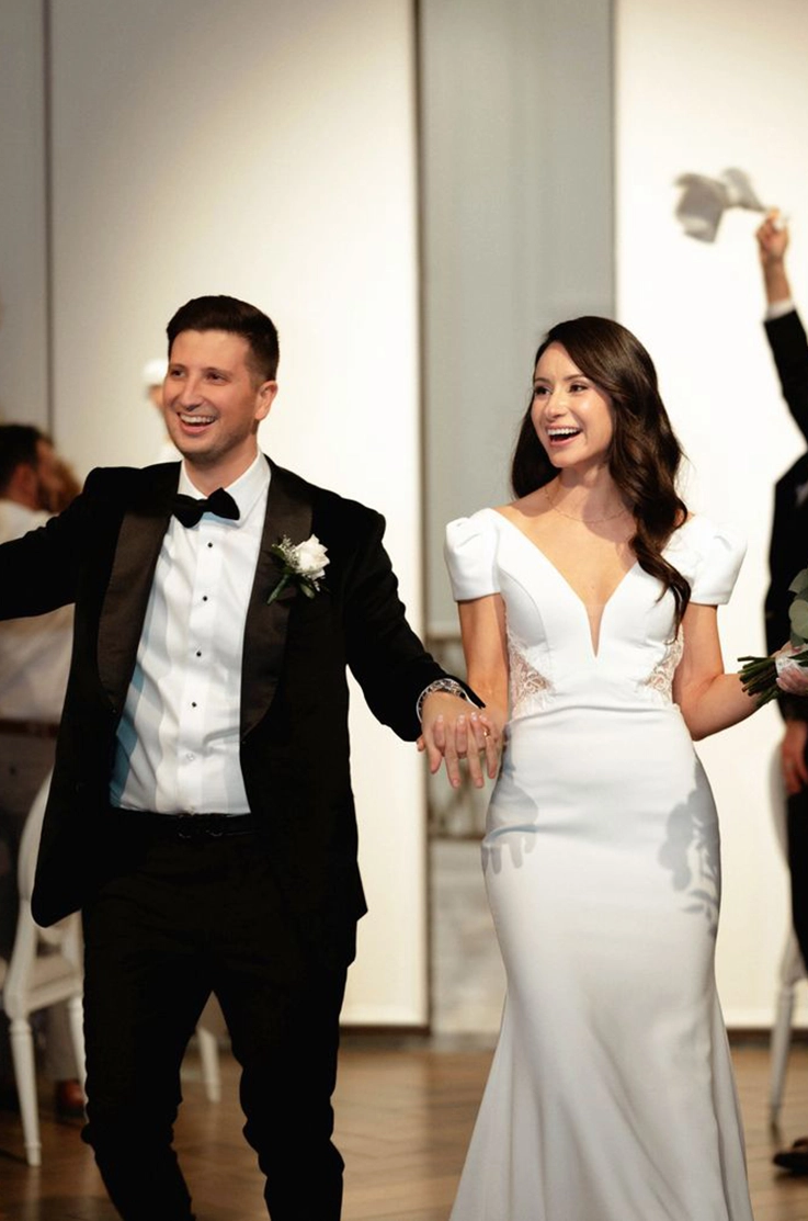 Bride in a fit and flare crepe gown with a V-neck and sleeves, holds hands with groom smiling as they walk down the aisle. 