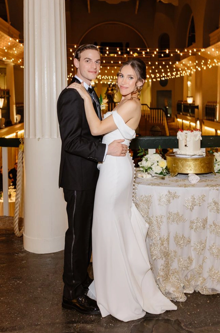 Groom embraces his bride in an off-shoulder fit and flare wedding dress with buttons at indoor venue with string lights.